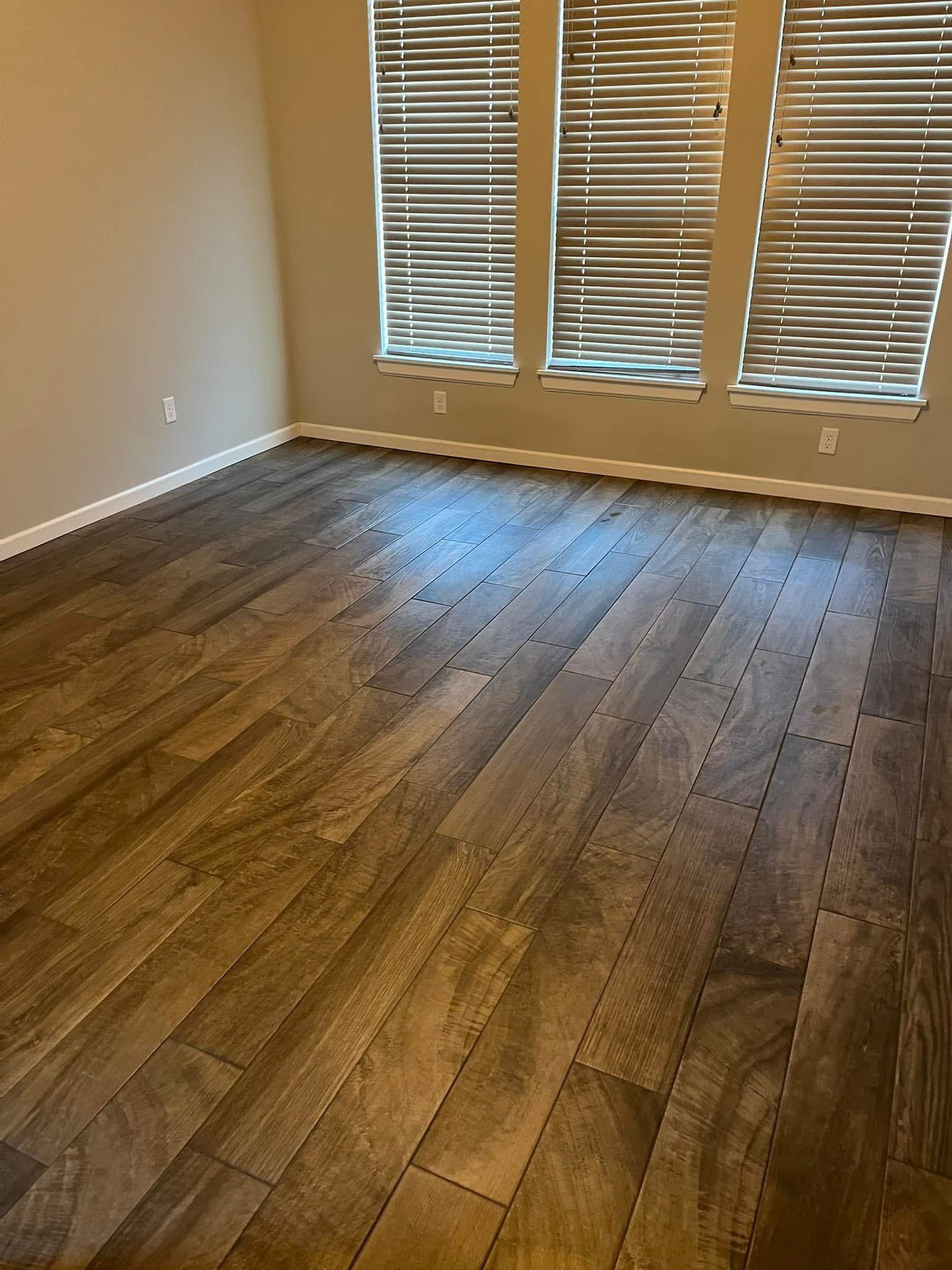 A room with wood-look tile flooring and three windows with closed blinds. Light beige walls.