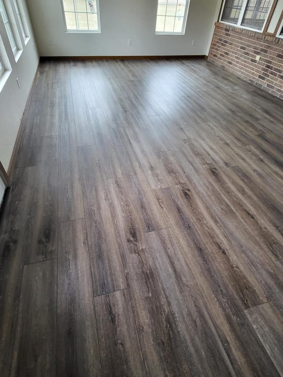 A room with wood-look flooring. Sunlight reflects off the dark brown and gray planks, and there is a brick wall visible
