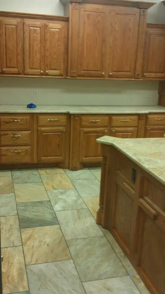 A kitchen with light wood cabinets, beige countertops, and a stone tile floor. An island is visible in the foreground.
