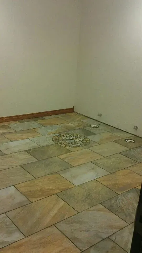 A corner of a room with tiled floor in tan and gray tones, featuring a decorative circular pattern. Light beige walls.