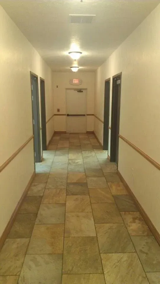Hallway with tiled floor, doors on both sides, and exit sign at the end. Beige walls and light fixtures.