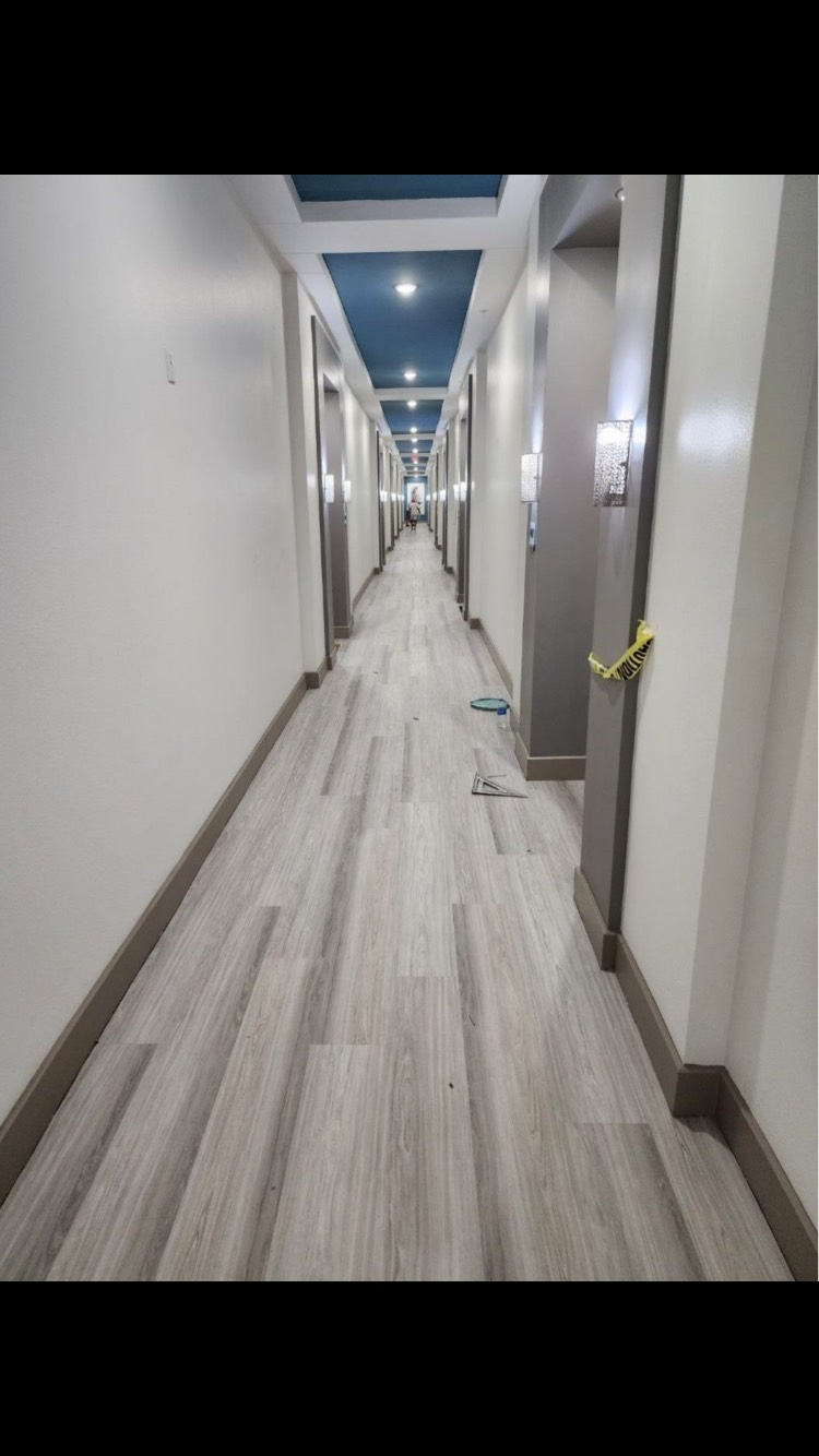 Long, empty hallway with light wood-look flooring and white walls, lined with closed doors. Blue accent ceiling.