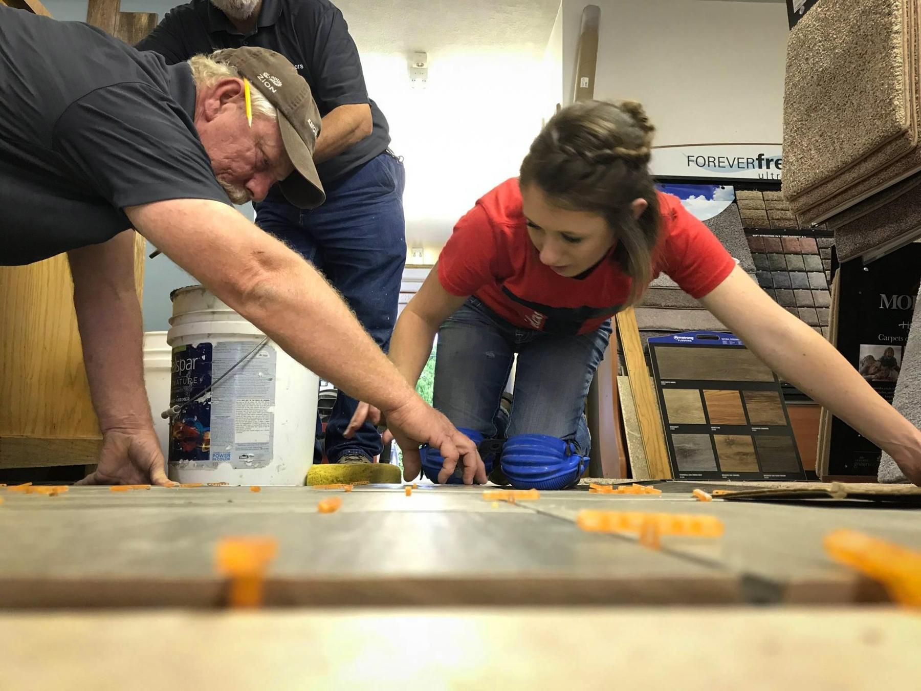 Three people installing floor tiles, with a woman kneeling and using orange leveling clips.