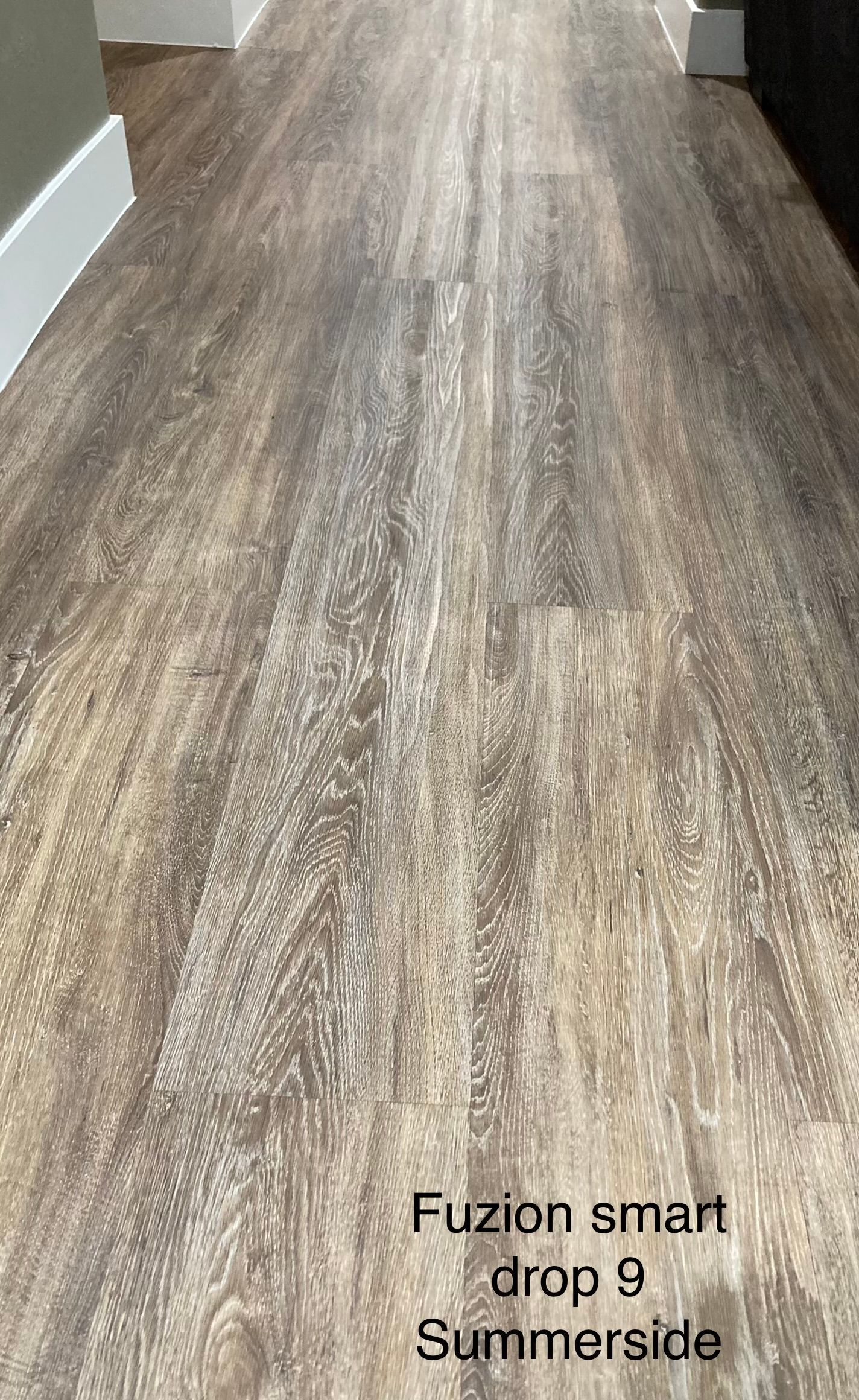 Wood-look flooring in a hallway with dark and light brown tones. White baseboards line the walls.