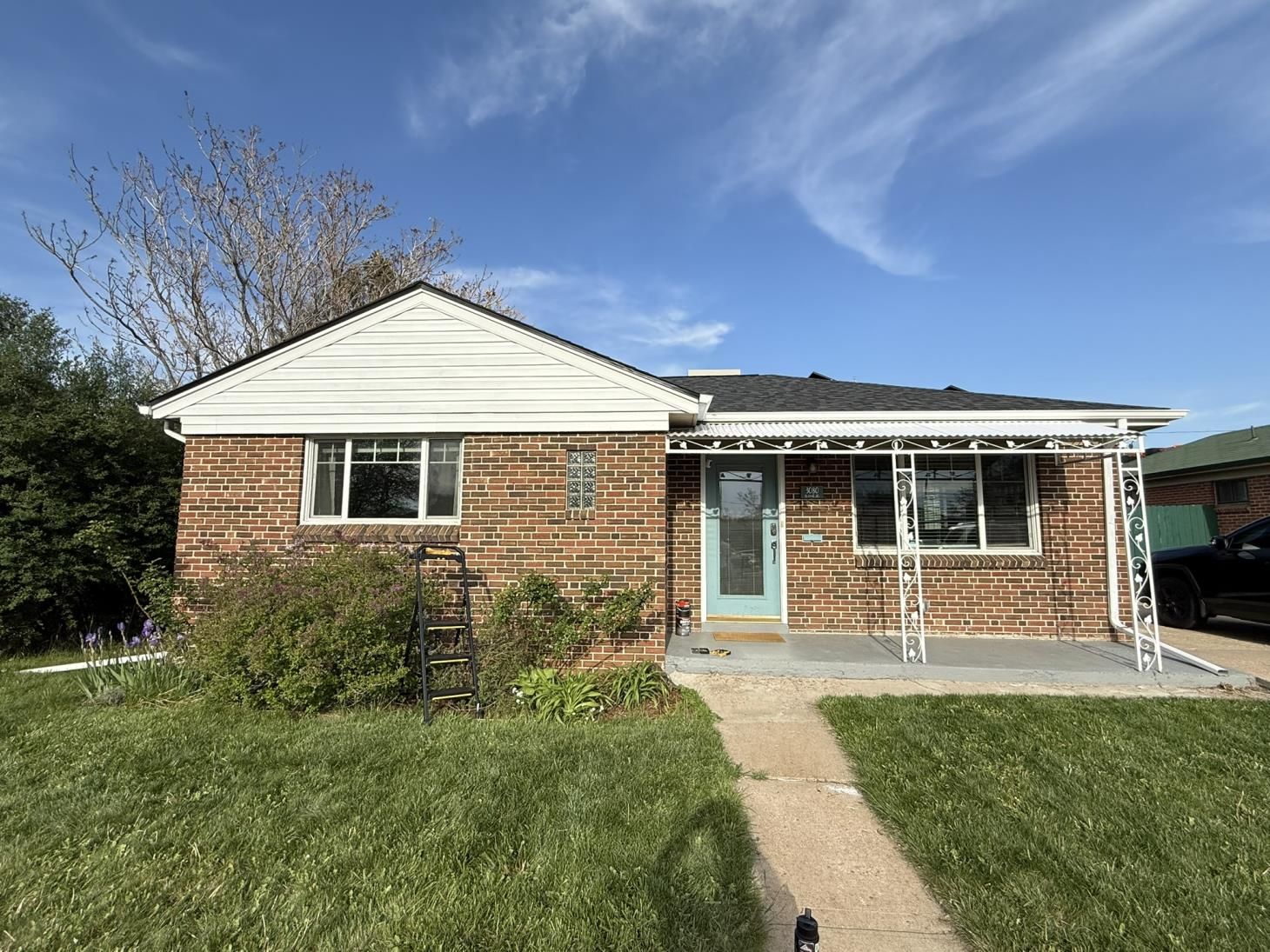 Brick house with blue door and white porch under a blue sky, overgrown yard.