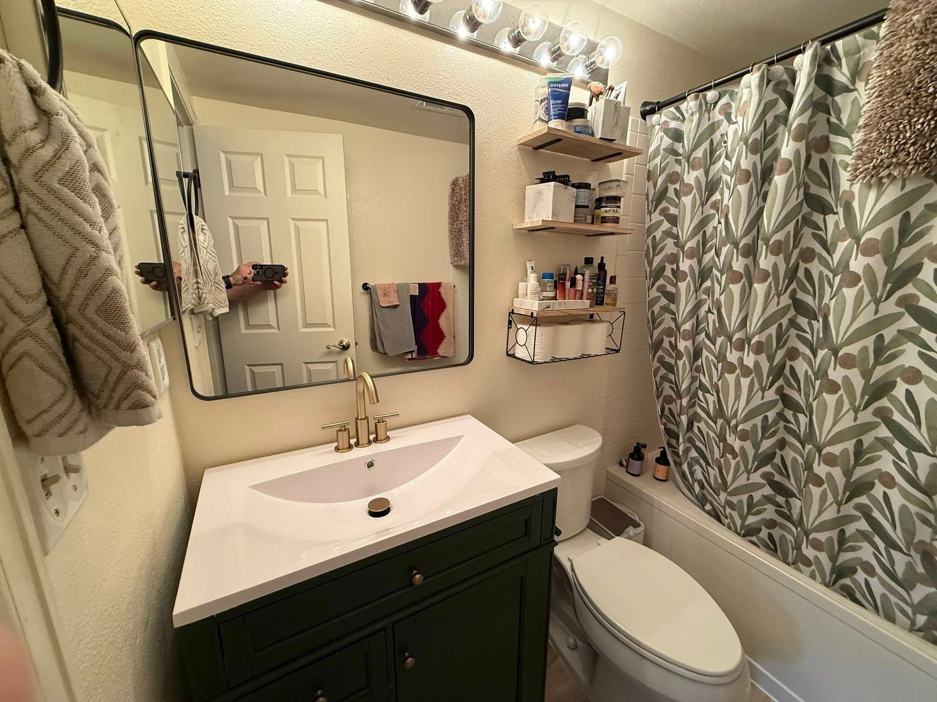 Bathroom with green vanity, black-framed mirror, shelves with toiletries, and a shower with olive leaf print curtain.