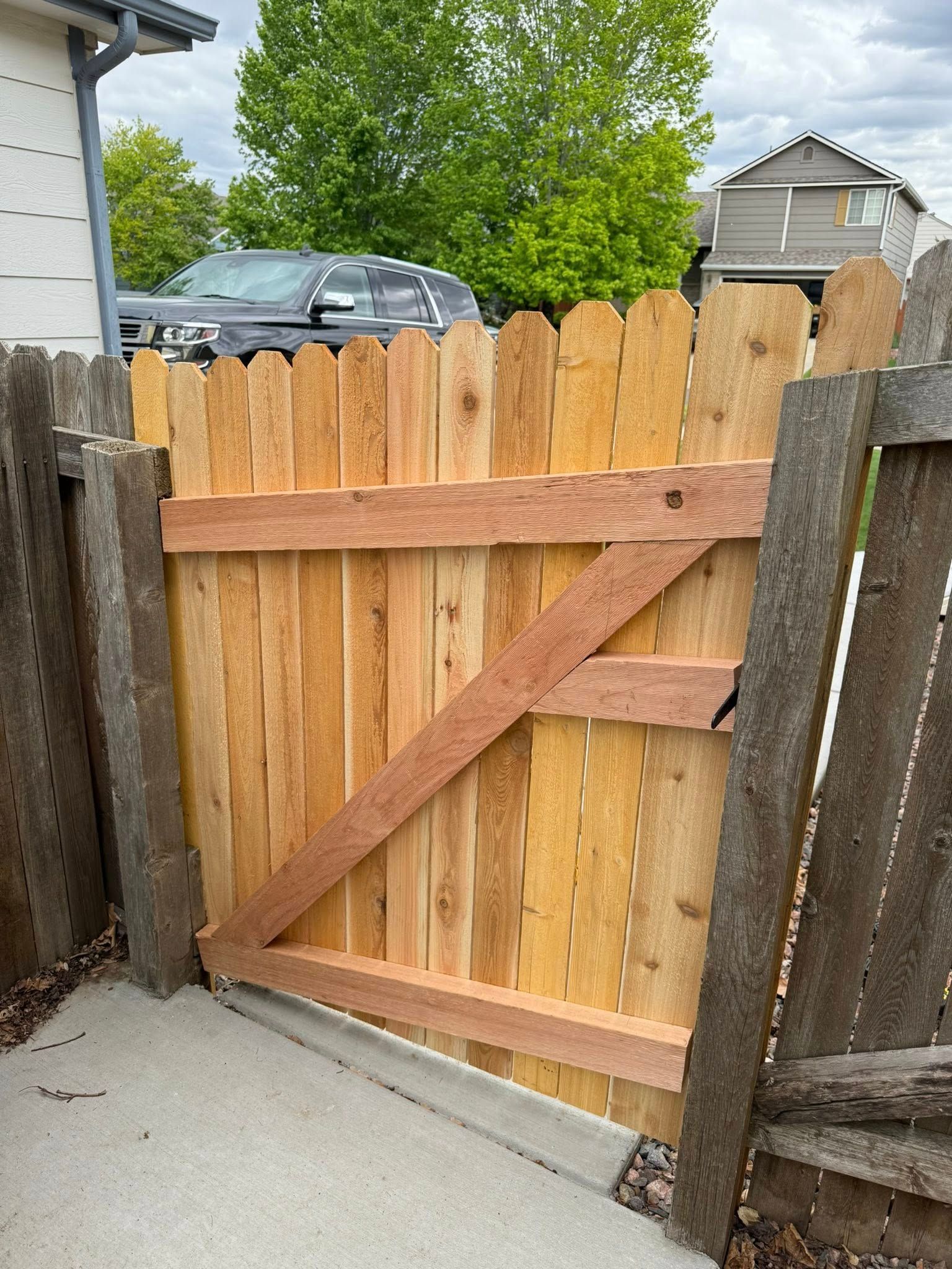 Wooden gate, light brown planks, diagonal brace, connects to fence posts, gray concrete ground.