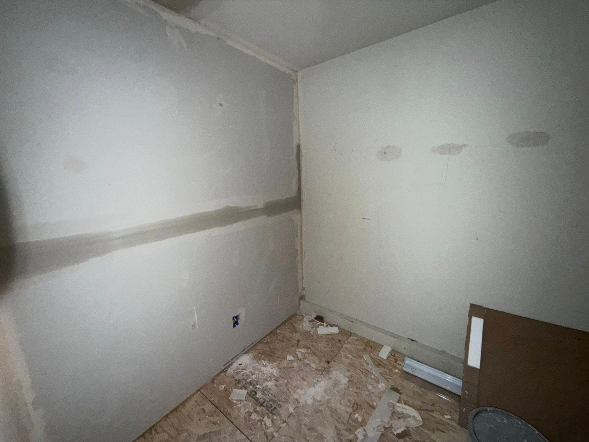 An unfinished room with drywall walls and a damaged wood floor.