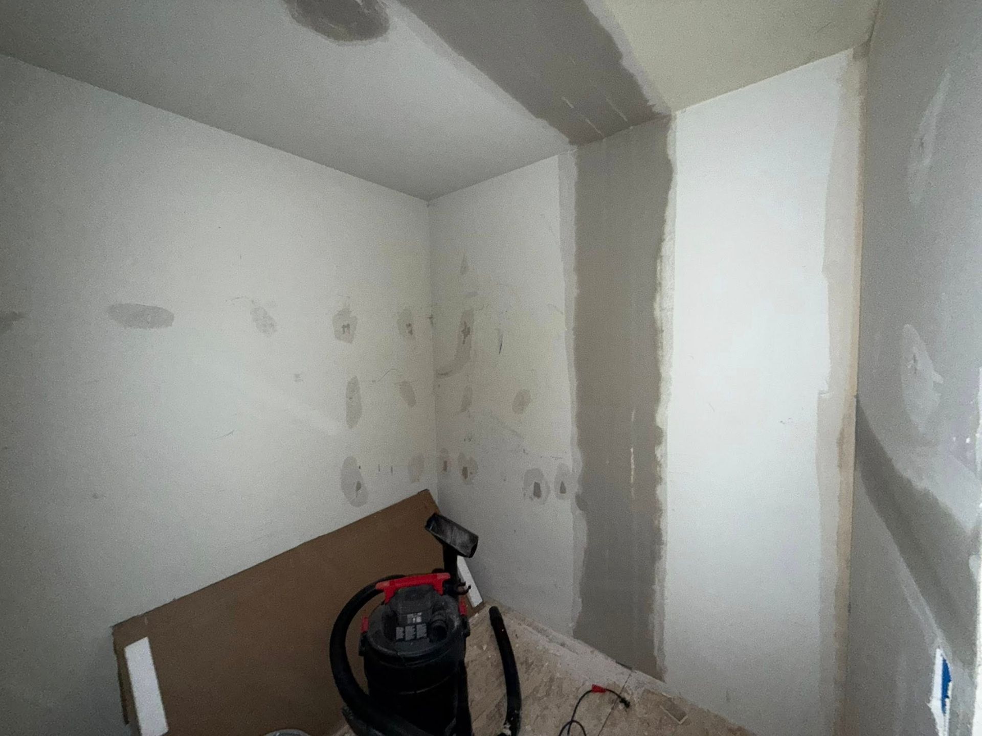 Interior corner of a room under construction with white walls, patching, and a vacuum cleaner.