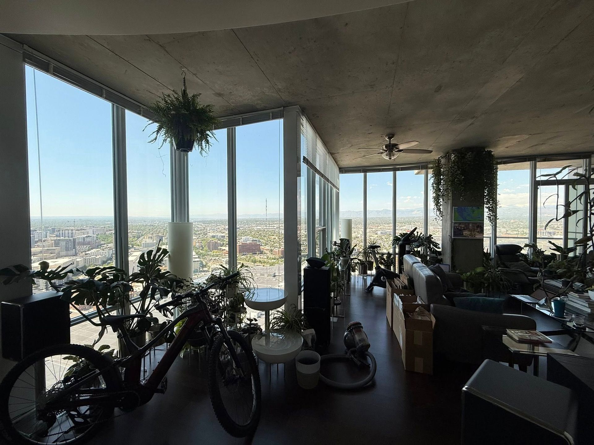 High-rise apartment interior with city view. Numerous plants, a bicycle, and various furnishings are present.