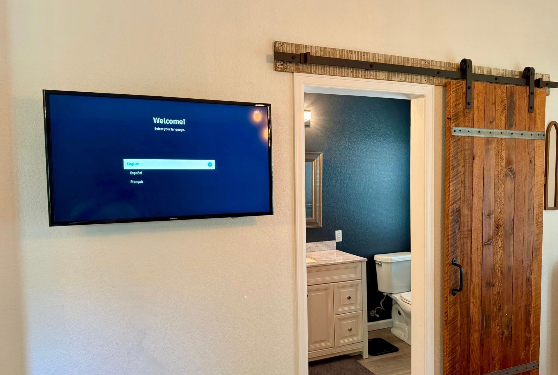 TV on a white wall, next to an open door with a rustic wooden sliding door, revealing a bathroom.