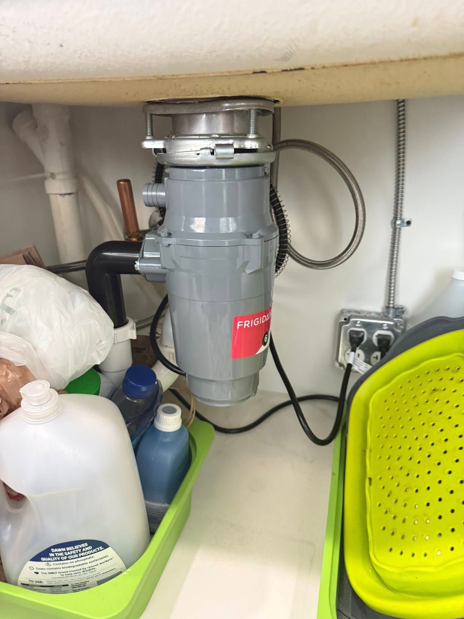 Garbage disposal under a kitchen sink. Grey disposal, black drainpipe, electrical outlet, and cleaning supplies.