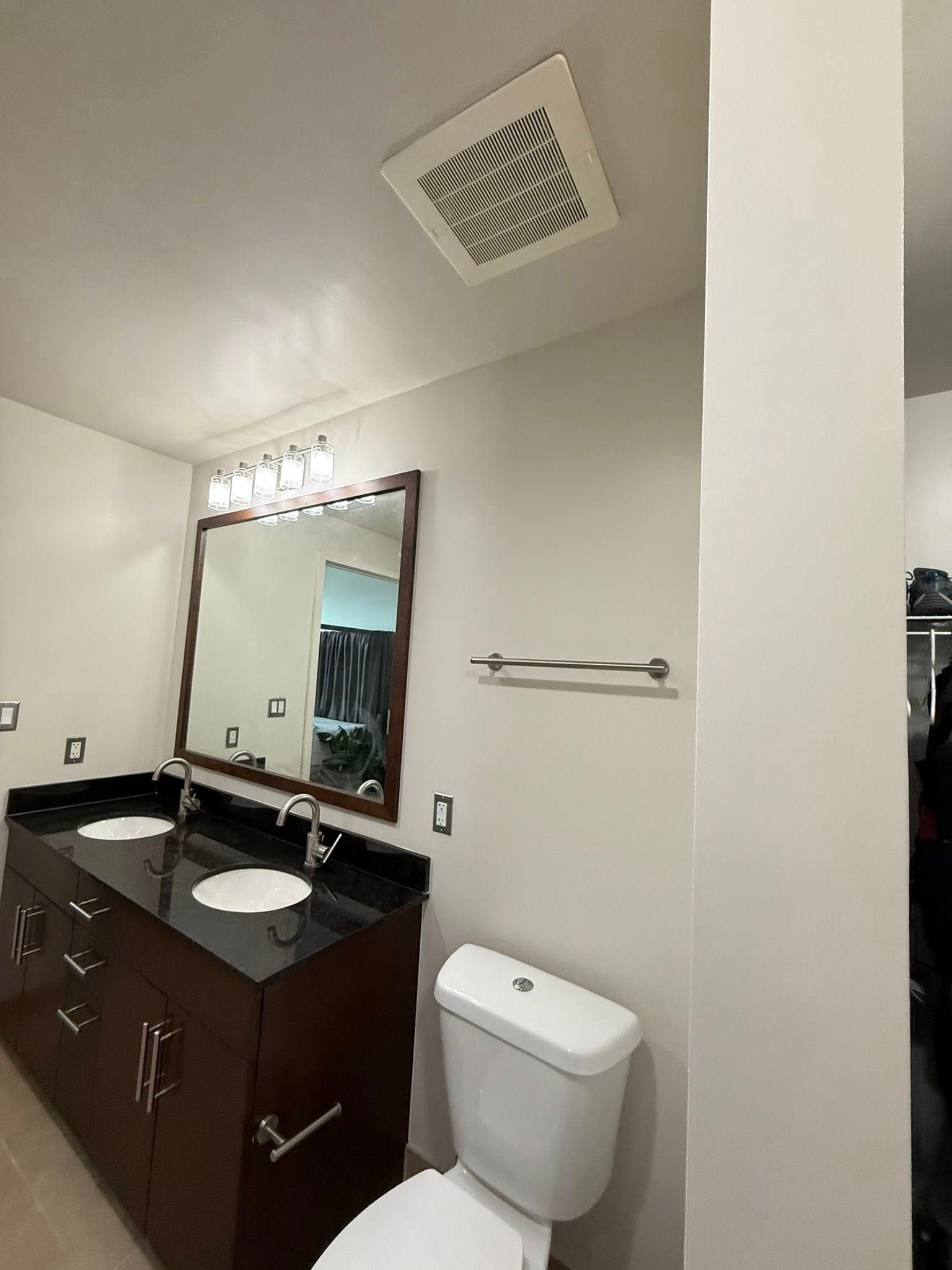 Bathroom with dark wood vanity, double sinks, a mirror, and a toilet. A ceiling vent and towel bar are visible.