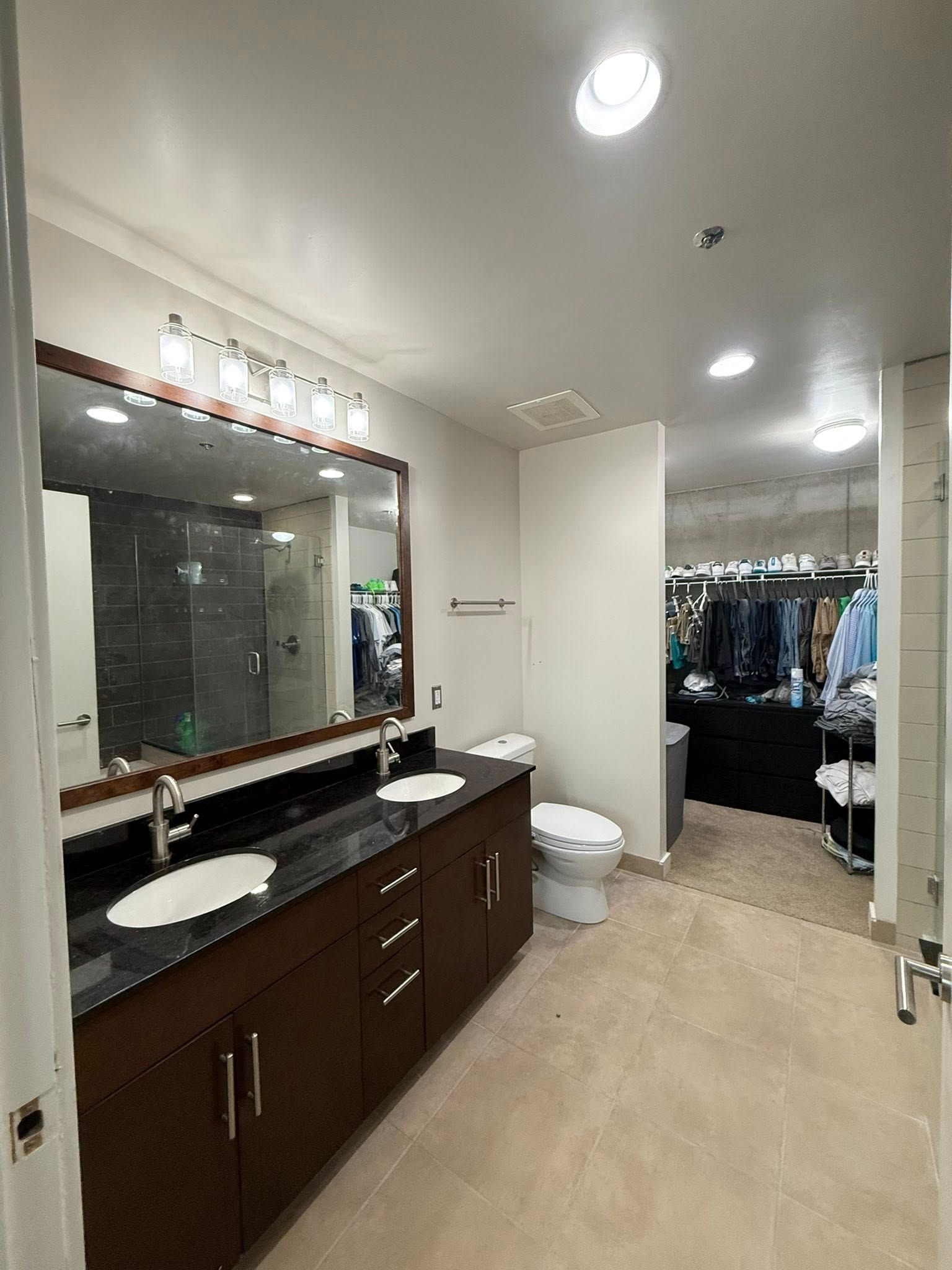 Bathroom with double vanity, toilet, and walk-in closet. Brown cabinets, dark countertop, and beige carpet.