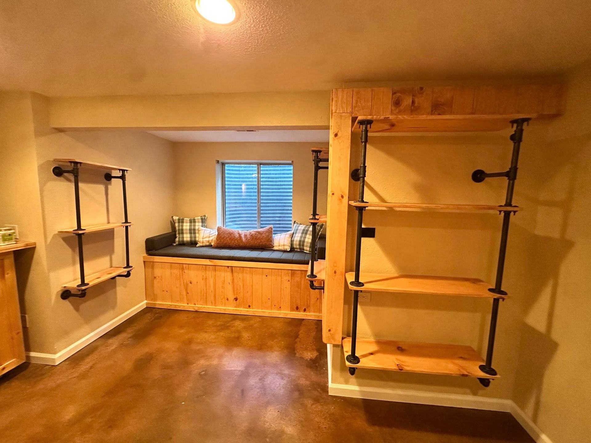 Cozy reading nook with built-in bench, window, and pipe shelving on wood and stained concrete floor.