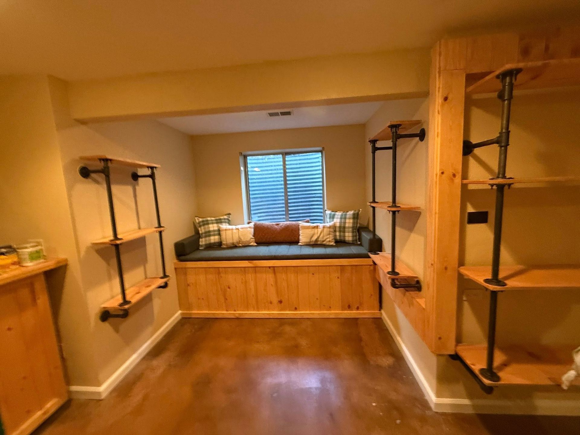 Cozy reading nook with built-in bench, window, and shelves made with wood and black pipes.