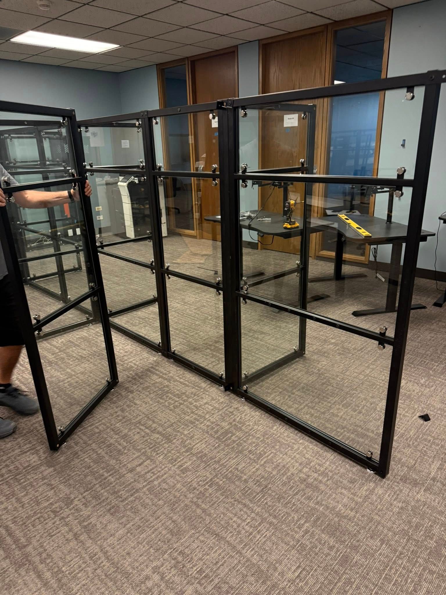 Person holding a black framed glass panel, near assembled panels in a room with carpet and windows.