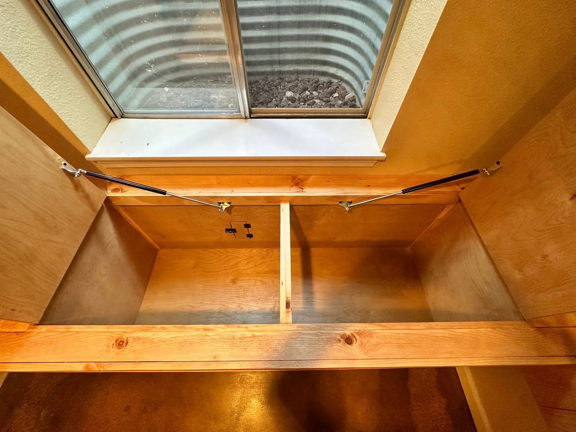 Interior view of a wooden storage unit built below a window. Two compartments and metal springs are visible.