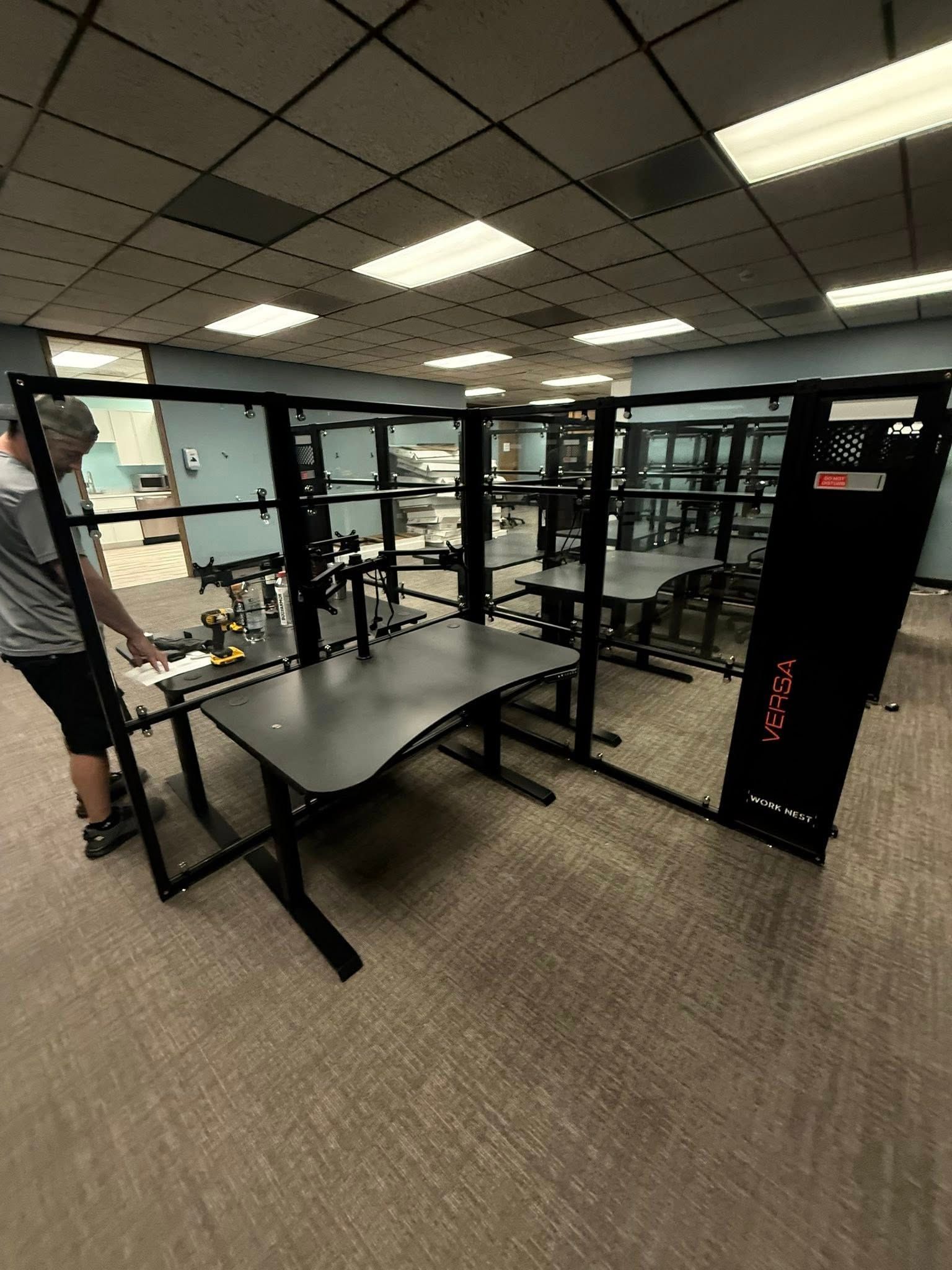 Person near a black desk inside a modular computer setup. The room has neutral carpet and ceiling lights.