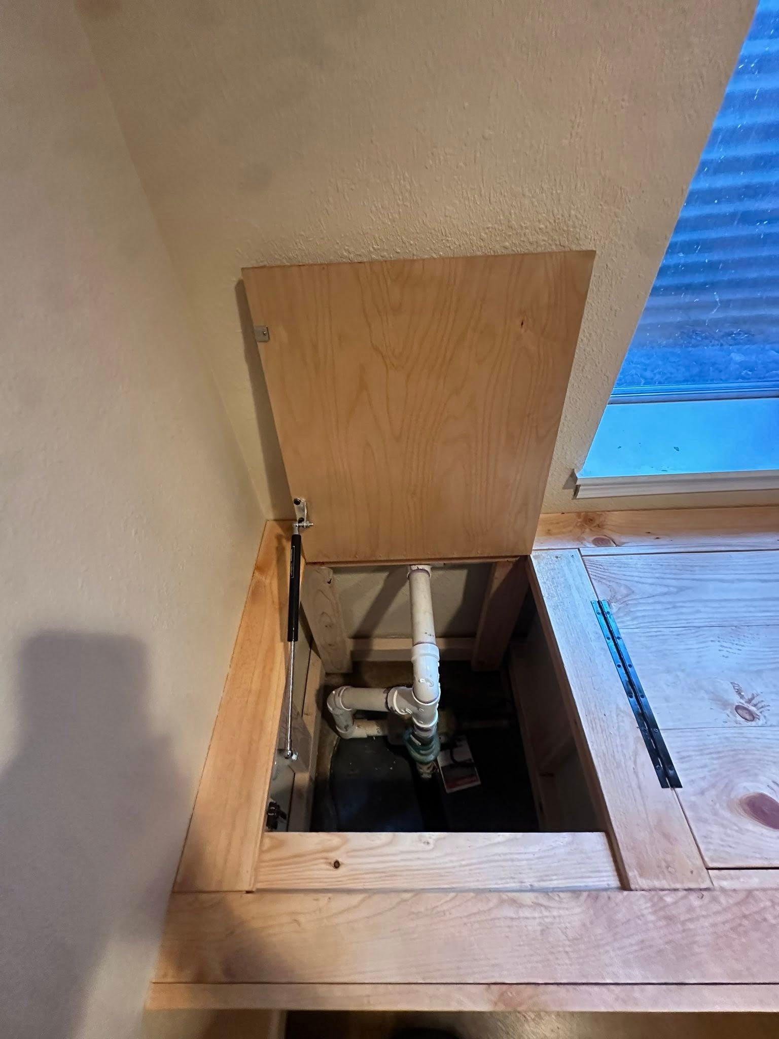 Hinged wooden access panel revealing plumbing in an enclosed space with a window.