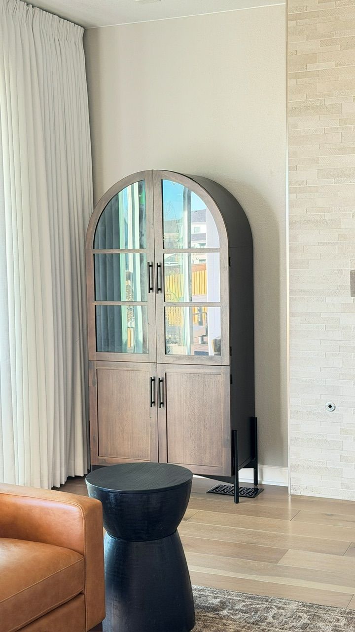 Wooden cabinet with arched top and glass doors, near a window, black stool in front.