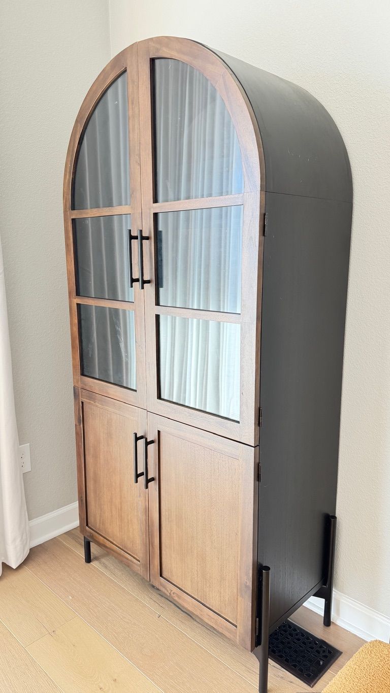 Black and wood-toned arched cabinet with glass door panels, on thin black legs, inside a room with light flooring.