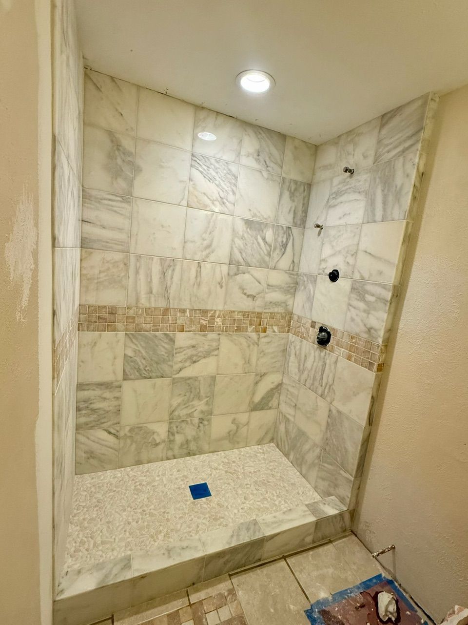 Newly tiled shower with marble-like walls and a pebble floor, featuring dark fixtures.