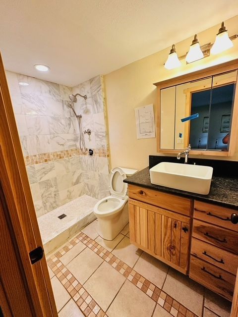 Bathroom with shower, toilet, vanity, and tan tile floor. Light wood cabinets, marble shower walls, and a white sink.