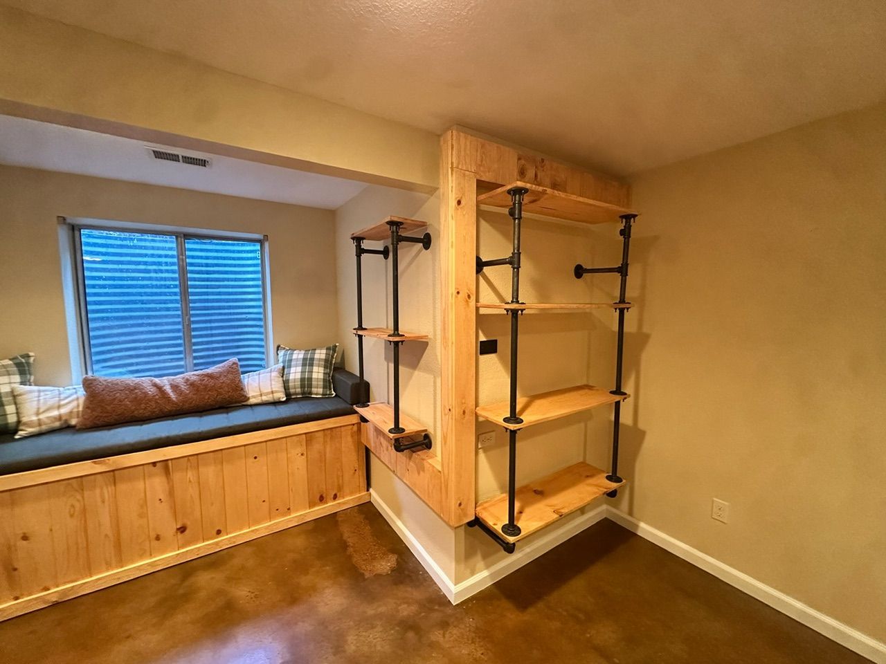 Wooden built-in storage bench by a window, with a pipe shelving unit in a room with stained floors.