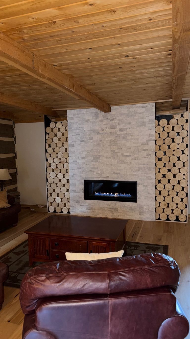 Cozy living room with fireplace and wood-stacked walls. Brown leather furniture, wood ceiling.