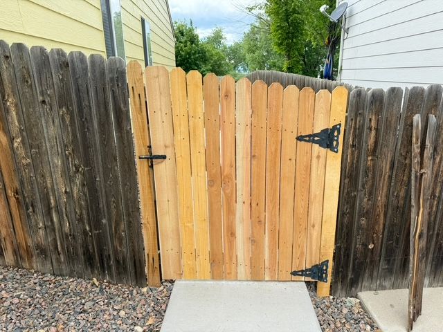 Wooden gate in a weathered fence, leading to a concrete walkway, set in a yard.