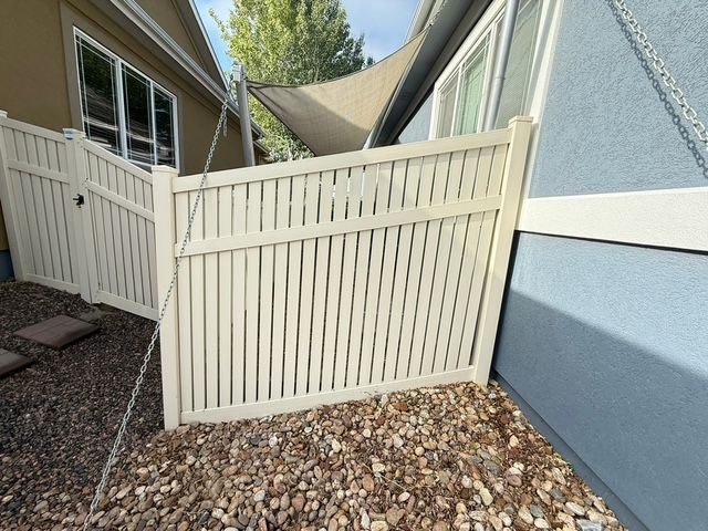 Beige vinyl fence between two houses, set in a gravel yard, with a shade sail in the background.