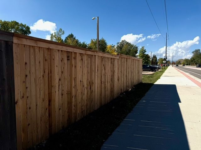 Wooden fence next to a sidewalk and road, under a blue sky with light clouds.