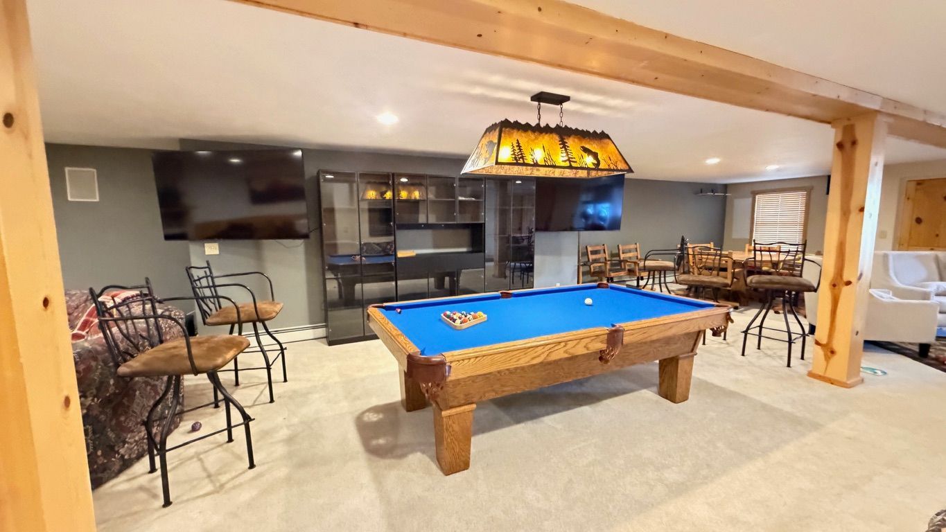 Basement recreation room with a pool table, bar, seating, and TVs. Light wood beams and neutral colors.