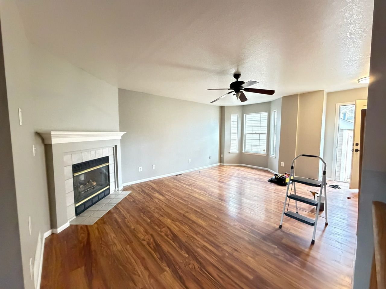 Living room with fireplace, wood floor, and gray walls. A ceiling fan, ladder, and window are present.