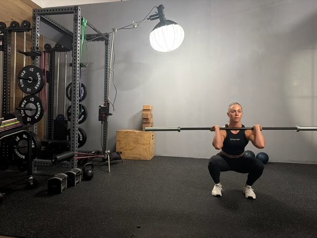 Woman performing a front squat with a barbell in a home gym. Dark hair, squatting position.