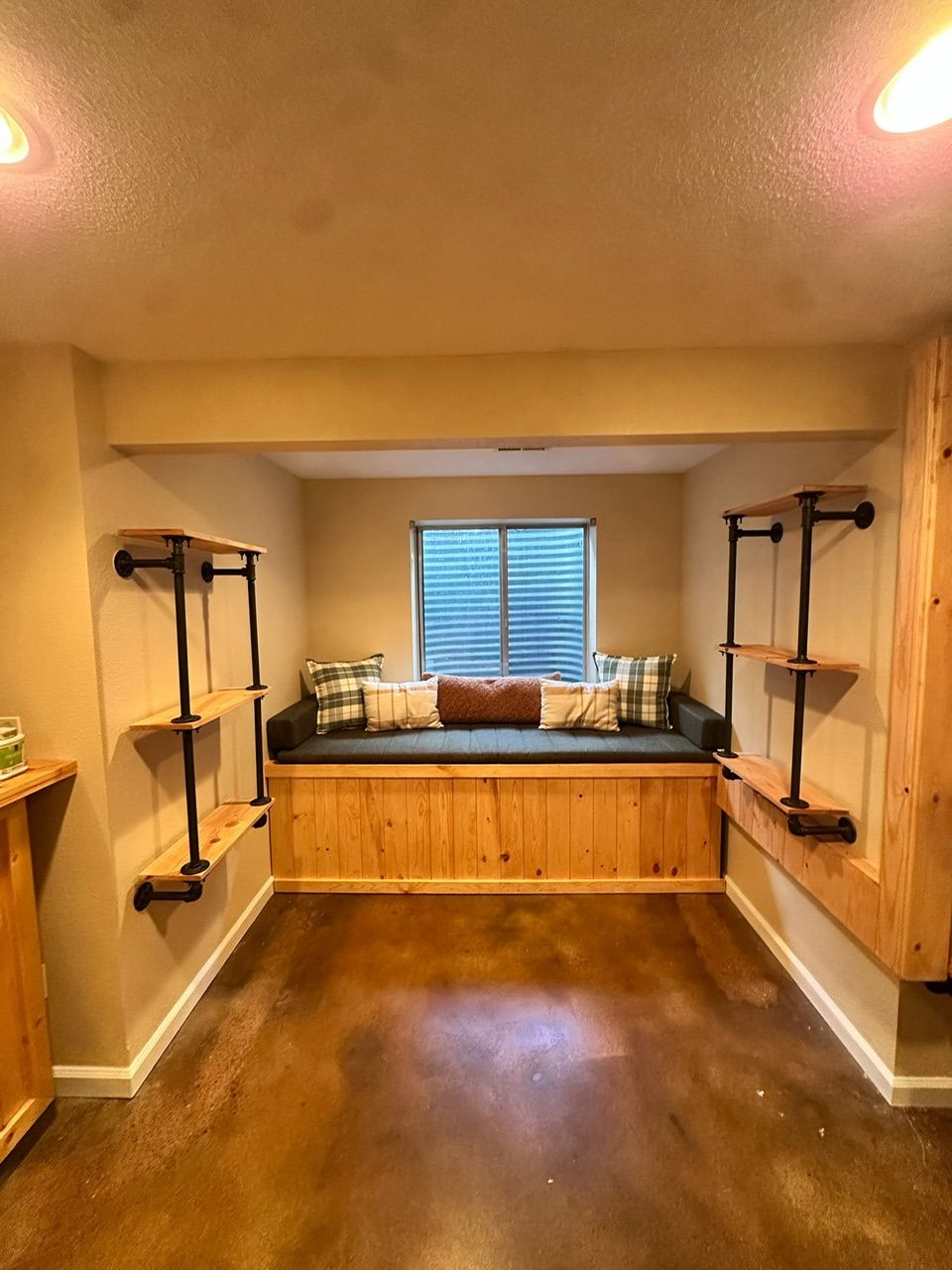 Cozy built-in bench seat with wood paneling, pillows, and window in a room with shelving and brown floors.