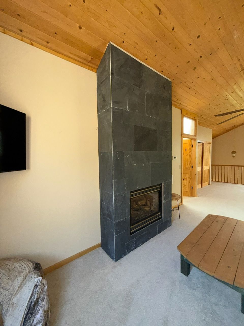 Stone fireplace in a living room with light carpet, wood ceiling, and a wooden table.