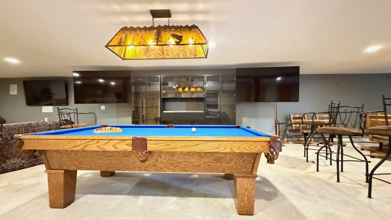 Pool table in a basement with bar stools, TVs, and decorative lighting.