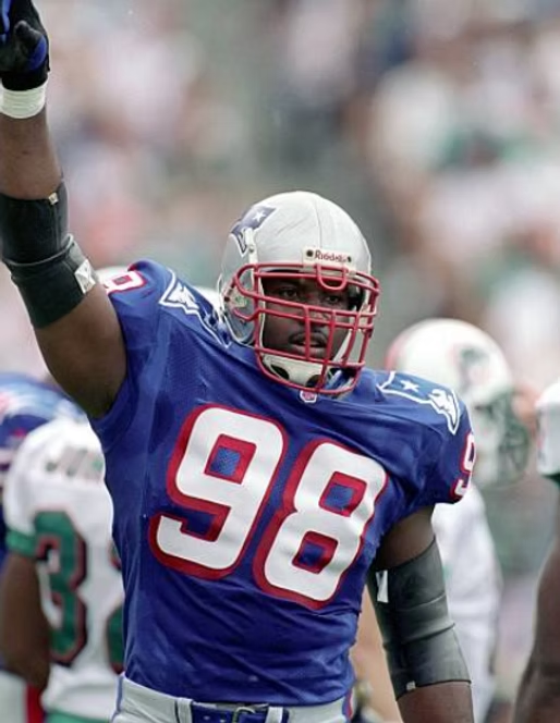 Football player in Patriots uniform, number 98, raises arm in stadium, blurred background.