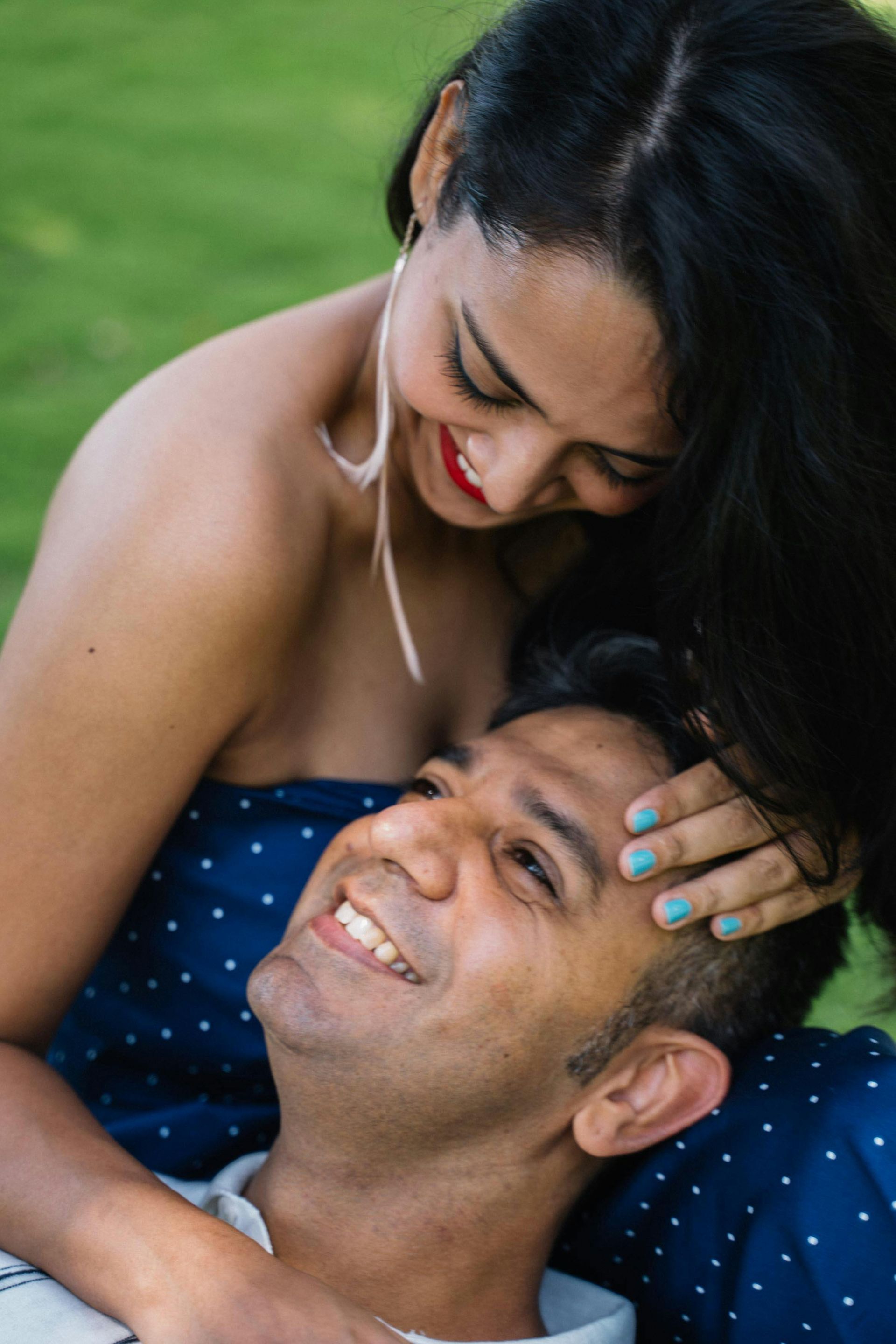 Woman smiling, head resting on man's head; both are outdoors in a grassy area.
