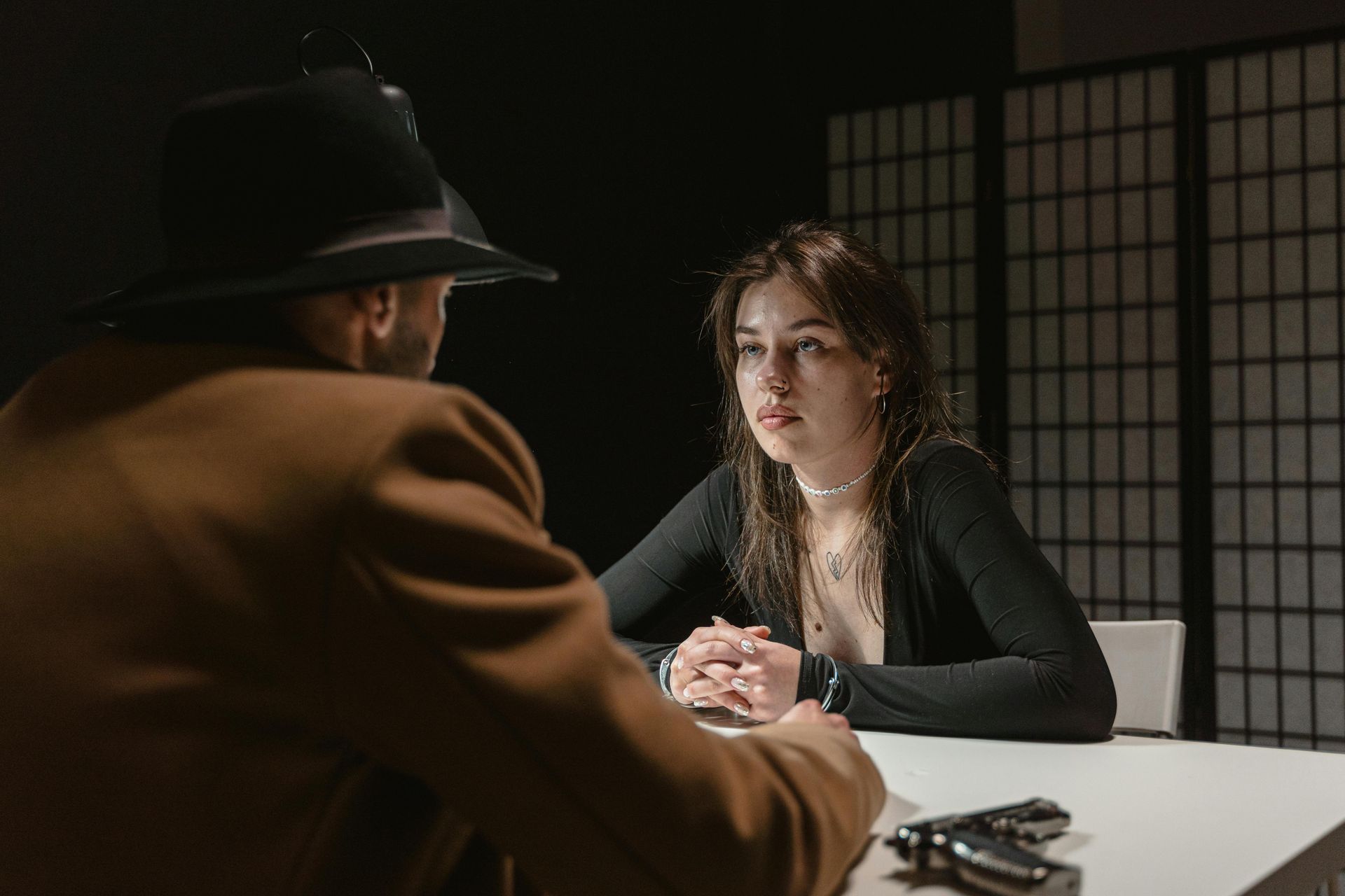 A person in a fedora and coat interviewing a person at a table, low lighting, gun visible.
