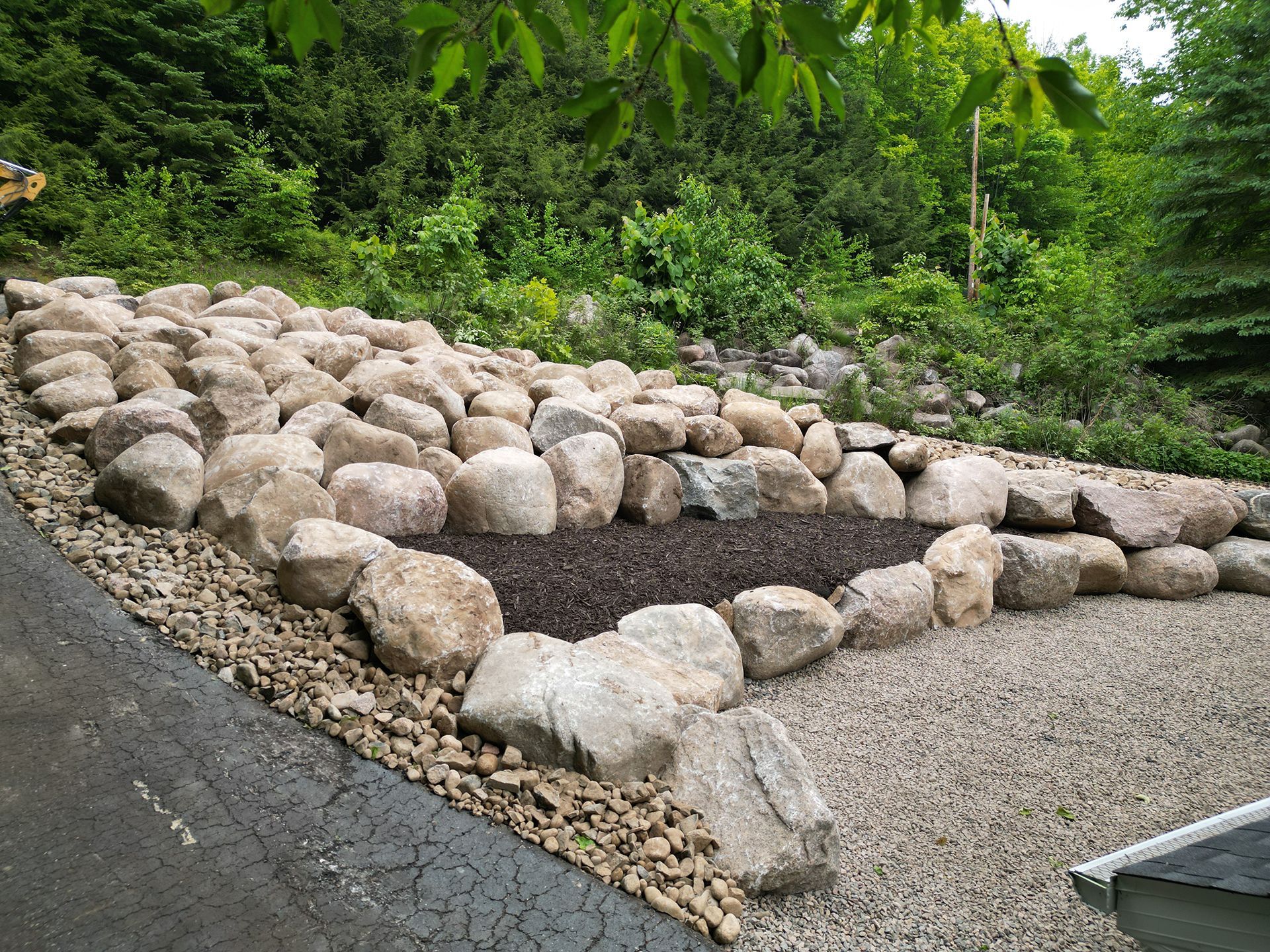 Rock garden with dark mulch bed, gravel path, and large rocks on a hillside with greenery in the background.