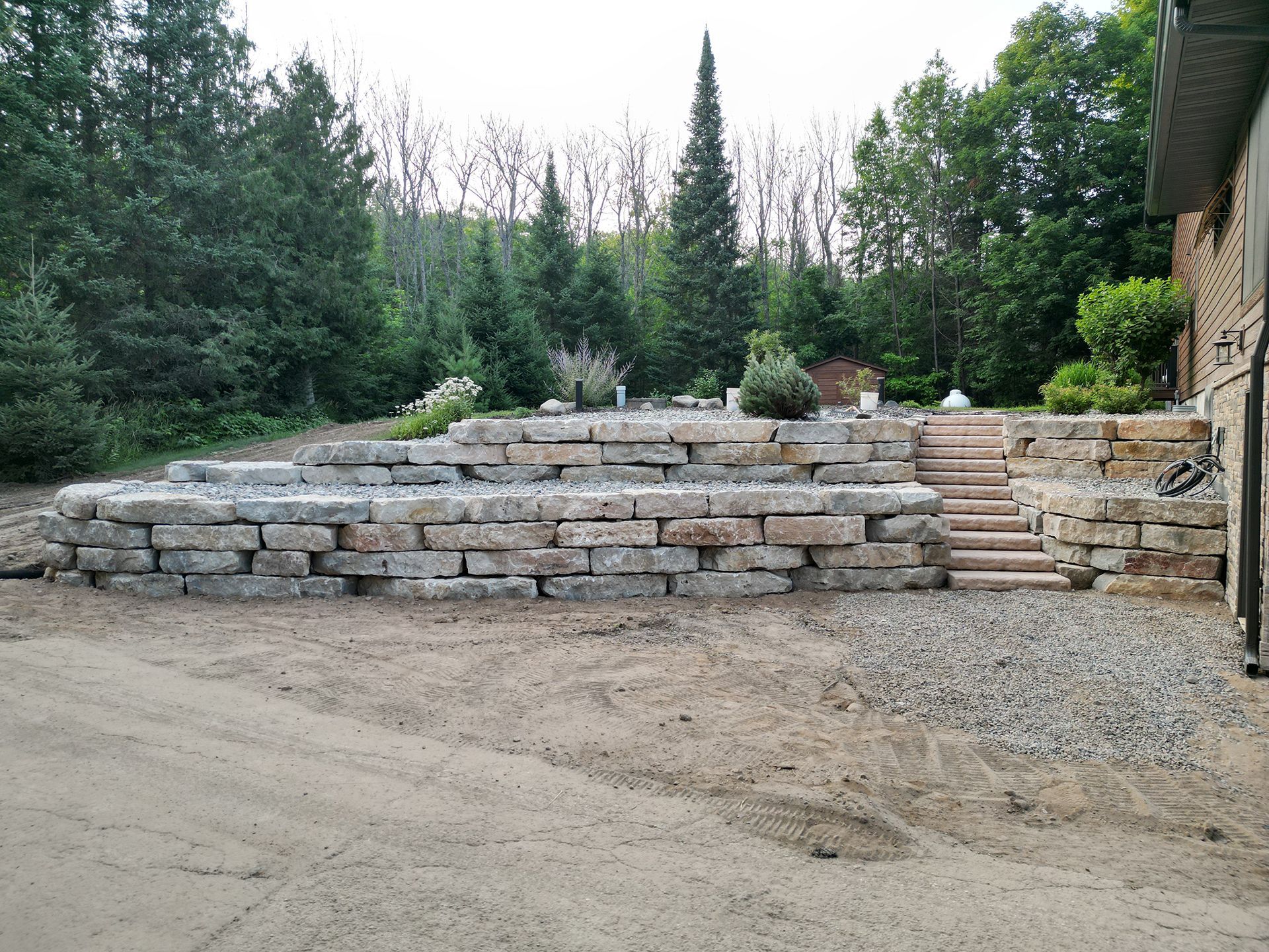 Stone retaining wall with steps, on a sloped yard. Trees and a small shed in the background.