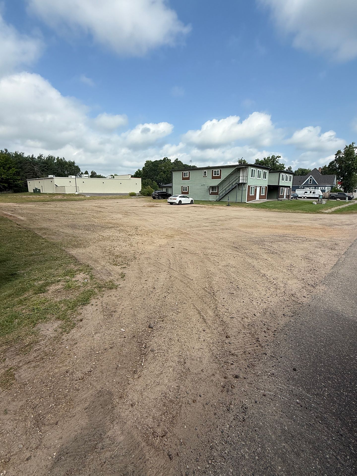 Dirt parking lot with two-story green building, white building, blue sky, and a car.