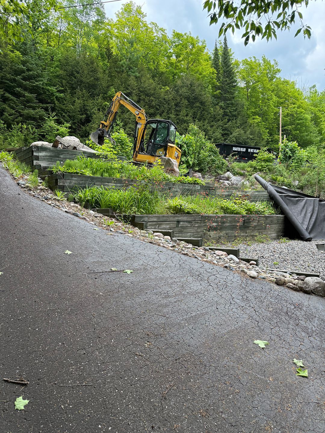 Yellow excavator on a hillside, working on a tiered landscape near a road and trees.