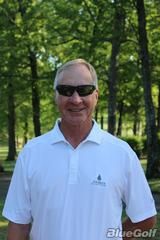 Tennis player in white shorts and visor swings racket on green court, shadow visible.