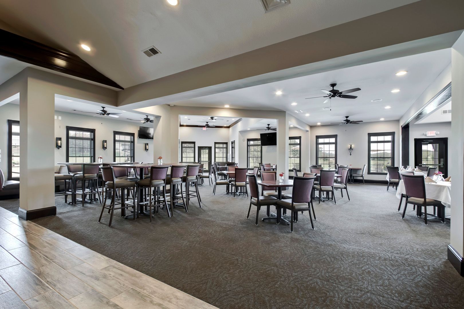Spacious, neutral-toned dining room with tables, chairs, and bar stools. Windows provide natural light.