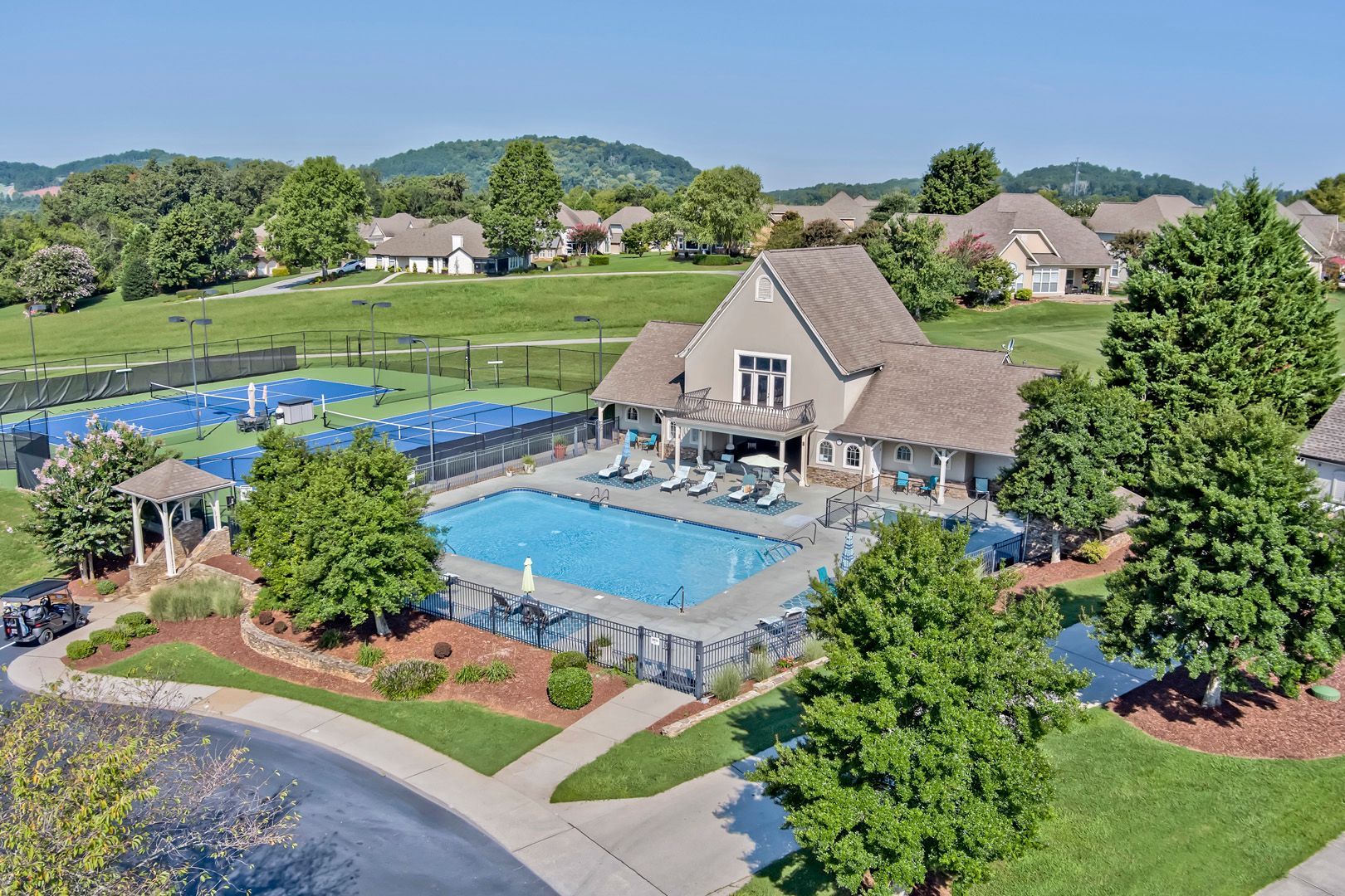Pool, tennis courts, and clubhouse in a residential area, surrounded by green lawns and trees under a clear blue sky.