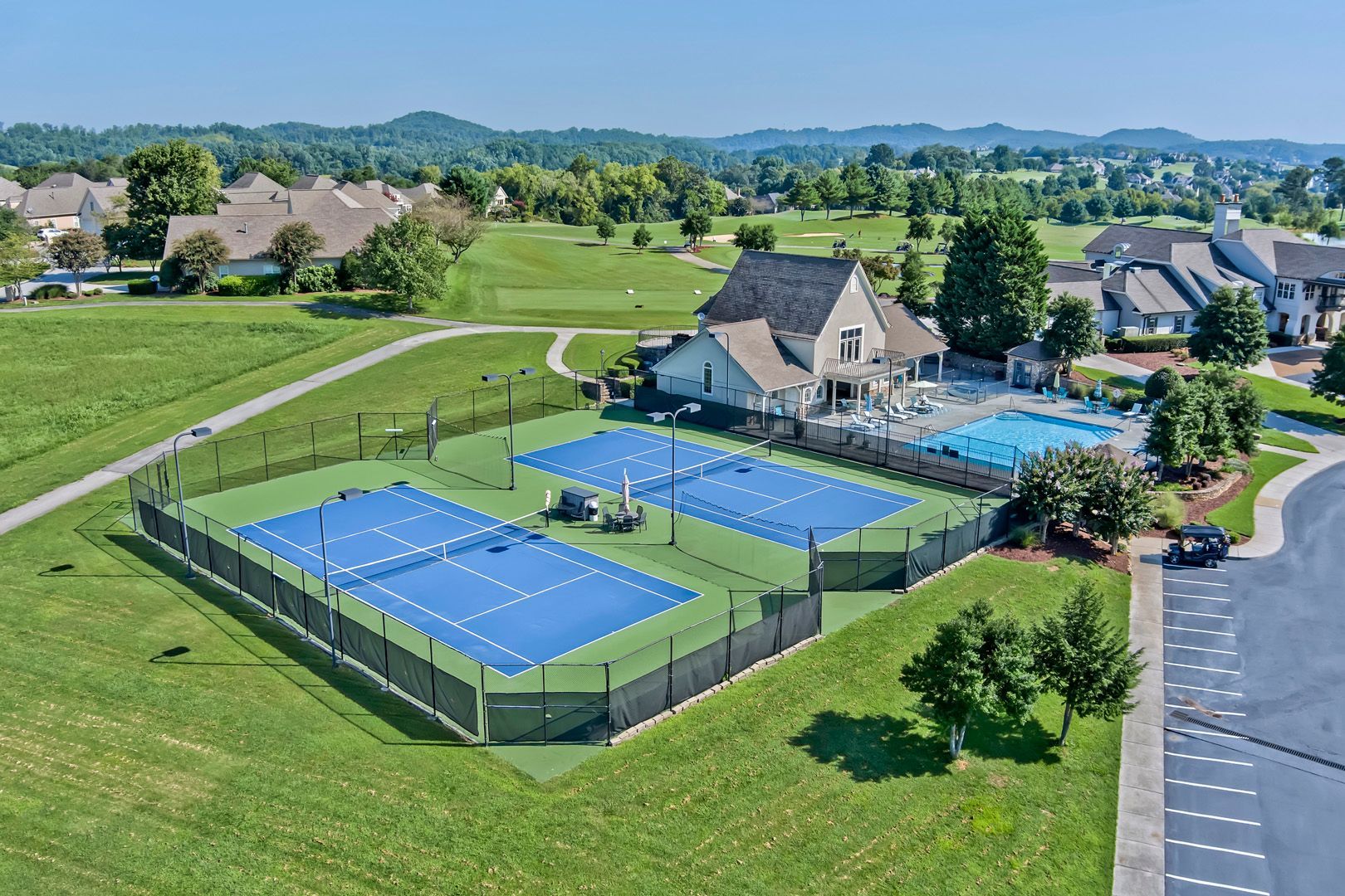 Aerial view of two blue tennis courts, a pool, and houses in a green landscape.